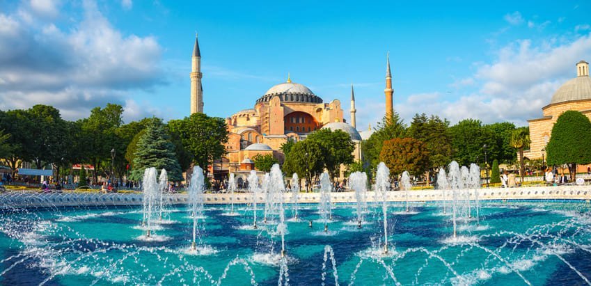 Бірюзовий фонтан перед Айя-Софією, оточений зеленню, купол і мінарети на тлі блакитного неба – спокійна, знакова панорама Стамбула.