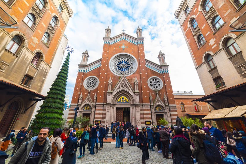 Величний фасад базиліки Святого Антонія з розетками й вежами на тлі блакитного неба, жвавий дворик із відвідувачами.