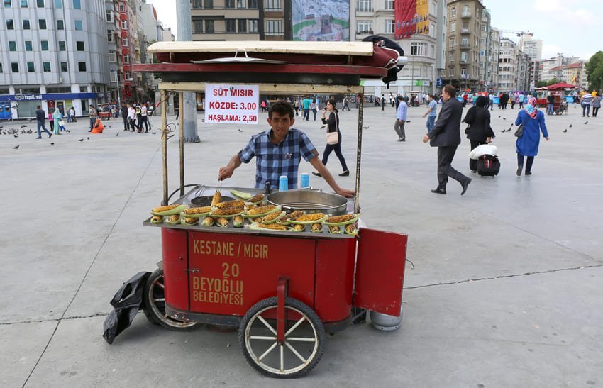 Вуличний продавець із червоним візком на великій міській площі пропонує варену та смажену кукурудзу, навколо рухлива юрба.