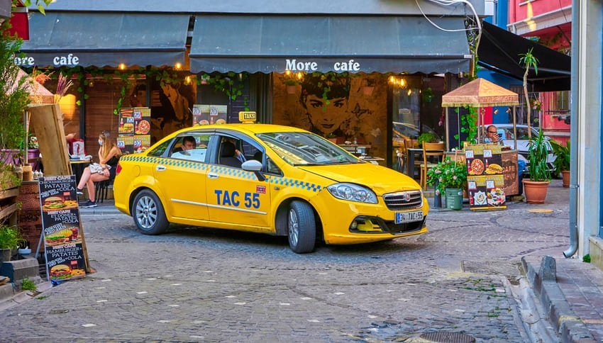 Жовте офіційне таксі з номером стоїть на вузькій вуличці біля фасаду кав’ярні з рослинами та крамницями в типовому турецькому місті.