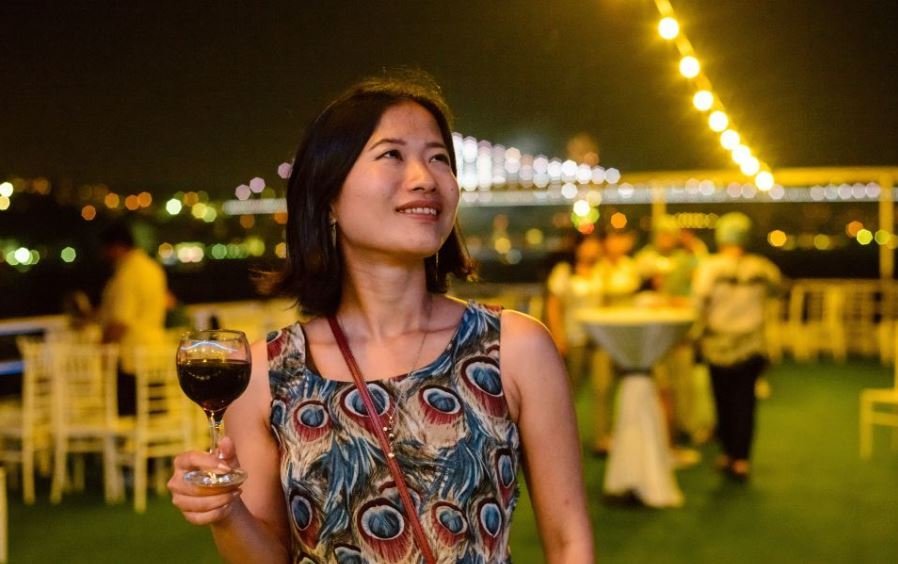 Жінка тримає келих вина на палубі човна, нічне освітлення, гірлянди вогників, гості на задньому плані та вид на міст або місто над Босфором.