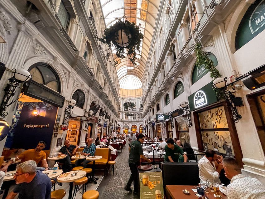 Критий пасаж із елегантними столами, скляним склепінням і витонченою архітектурою, наповнений теплим світлом та відвідувачами.
