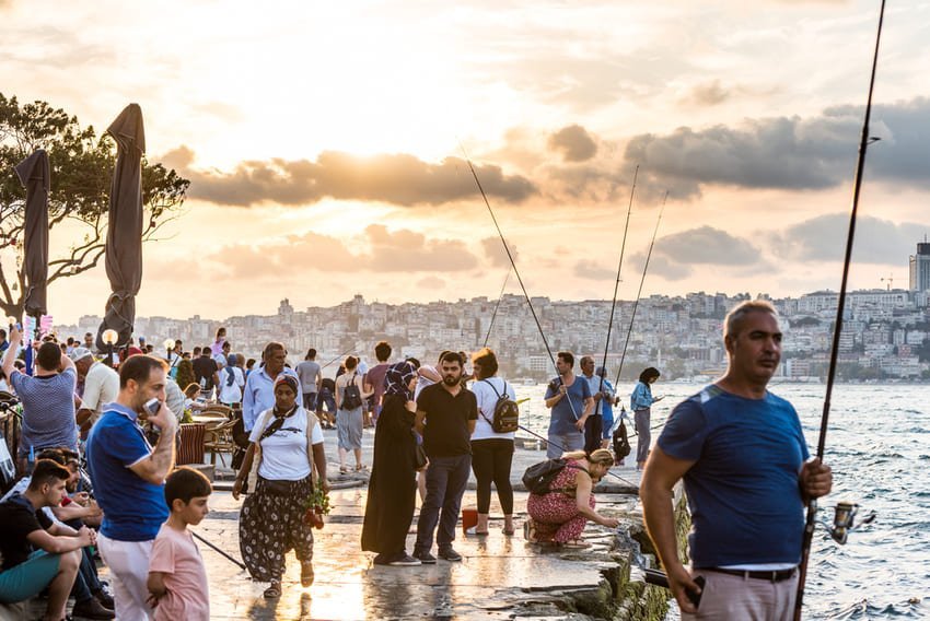 Набережна в променях заходу сонця, рибалки й сім’ї на фоні міської панорами, вудки, золоте світло й дружня атмосфера біля води.
