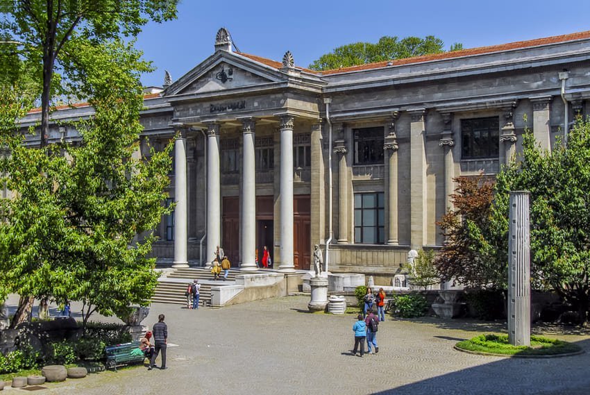 Неокласичний фасад археологічного музею Стамбула з колонами, фронтоном і деревами на передньому плані.