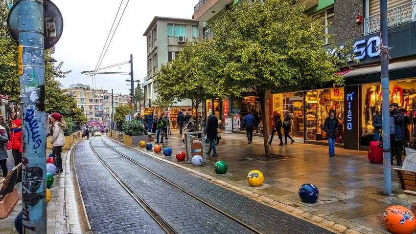 Пішохідна вулиця в Moda з магазинами, кав’ярнями та деревами, прикрашена кольоровими кулями над рейками трамваю.
