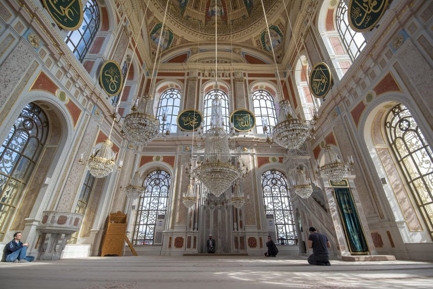 Я знаходжусь всередині мечеті Ортакьой у Стамбулі, з великими вікнами, кришталевими люстрами, написами арабською та розписаним куполом.