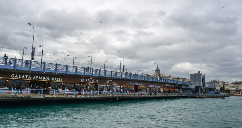 Я на човні перед мостом Галата, під яким розташовані рибні ресторани, а на горизонті видніється вежа Галата під сірим небом.